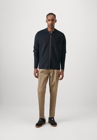 Navy zip-up sweater with a collar, front pocket, beige pants, and black sneakers with gum soles, on a person standing against a plain backdrop.
