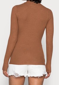 Osoba w dopasowanej brązowej sweterkowej bluzce z długim rękawem i białych zniszczonych szortach dżinsowych, widziana z tyłu.