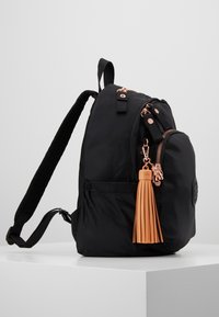 Schwarzer Rucksack mit gepolsterten Trägern, mehreren Reißverschlussfächern, Roségoldbeschlägen und einem hellbraunen Quasten-Schlüsselanhänger, der am Reißverschluss der Vordertasche hängt.