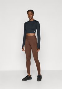 Strak aansluitend crop top met lange mouwen in donkerblauw, gecombineerd met een high-waisted bruine legging. Model draagt zwarte sportschoenen. Glad stoffen textuur.