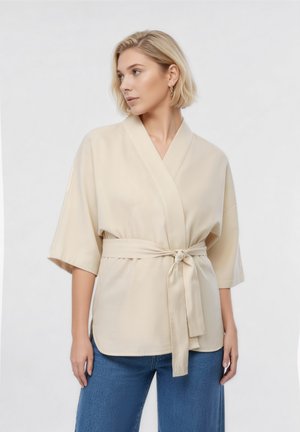 Vrouw met kort blond haar die een beige kimono-achtig top met ceintuur en een blauwe spijkerbroek draagt, kijkt naar links tegen een effen achtergrond.