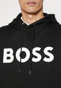Black hooded sweatshirt with prominent white "BOSS" text. Features drawstrings with silver tips and a soft, textured fabric.