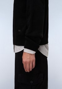 Maglione nero a maglia con polsini a coste, indossato sopra una camicia a righe leggera. L'outfit include pantaloni cargo neri con tasca frontale.
