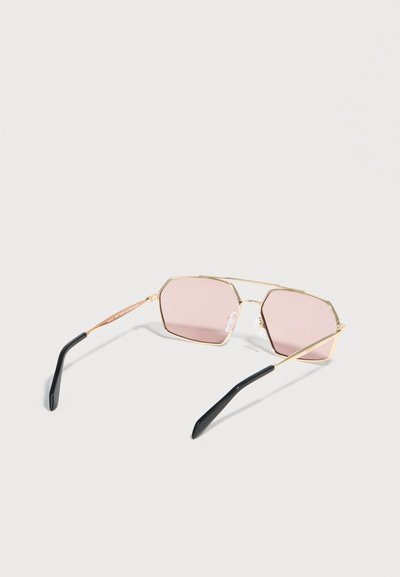 Γυαλιά ηλίου με χρυσή κορνίζα, γεωμετρικούς ροζ φακούς και μαύρα άκρα στο πλαίσιο, τοποθετημένα σε ανοιχτή επιφάνεια.