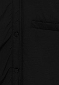 Gros plan sur le devant d'une chemise en tissu noir avec deux boutons-pression noirs et une poche poitrine horizontale du côté droit.