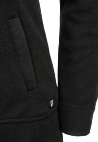 Črna flis jakna z bočnim žepom in majhno značko z logotipom. Tkanina je videti mehka z teksturirano površino in sproščenim krojem.