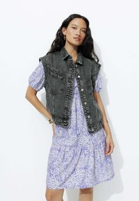 Veste en denim gris foncé avec des coutures d'épaule et des poches avant, superposée sur une robe légère violette à motif tacheté.