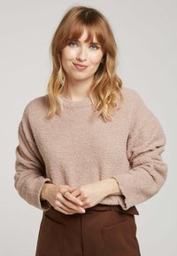Pull en rose clair, texturé, avec un col rond et une coupe décontractée. Manches longues avec des bords bruts. Associé à un pantalon marron taille haute.