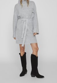 Abito grigio oversize con cintura a cravatta e maniche lunghe, abbinato a stivali neri in suede fino al ginocchio con tacco a blocco.