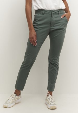 Persoon draagt een slim fit olijfgroene broek, witte sneakers en een wit ingestopt topje, staand met één hand in de zak.