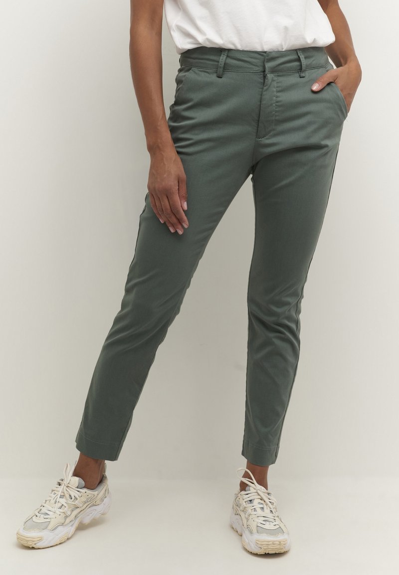 Personne portant un pantalon slim vert olive, des baskets blanches et un haut blanc rentré, debout avec une main dans la poche.