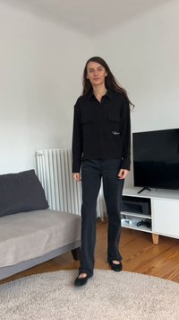 Juoda marškiniai su sagomis ir dviem krūtinės kišenėmis, derinami su tamsiais džinsais ir juodais batais. Lygi tekstūra ir pritaikyta forma.