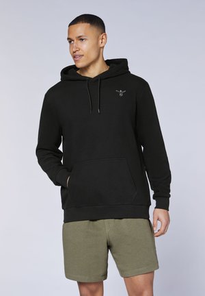 Mann trägt schwarzen Hoodie mit Kängurutasche und kleinem Logo, kombiniert mit olivgrünen Shorts, stehend vor schlichtem Hintergrund.