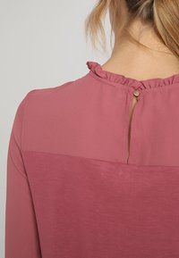 Blus med lång ärm i mjukt mauve tyg, med en volangkrage och en nyckelhålsknäppning med en dekorativ knapp bak.