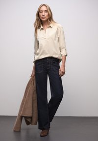 Jasnobeżowa koszula w paski z długim rękawem, ciemnoniebieskie dżinsy o szerokich nogawkach oraz brązowe zamszowe botki za kostkę; trzyma brązowy płaszcz z sztucznego futra.