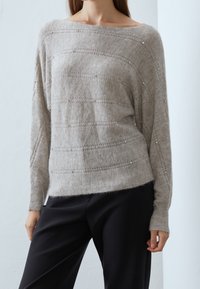 Femme portant un pull en maille gris clair orné de petites perles, associé à un pantalon noir ample, debout devant un fond uni clair.