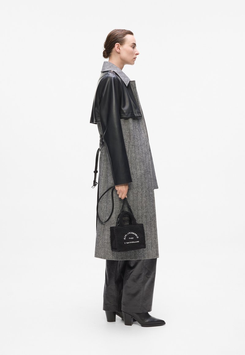Long manteau en mélange de laine gris avec des manches en cuir, associé à un pantalon en cuir noir brillant et un petit sac noir avec un texte blanc.