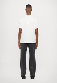 Hvid bomulds t-shirt med et lille label på ryggen, parret med løstsiddende mørke grå sweatpants med rød tekst på venstre ben.