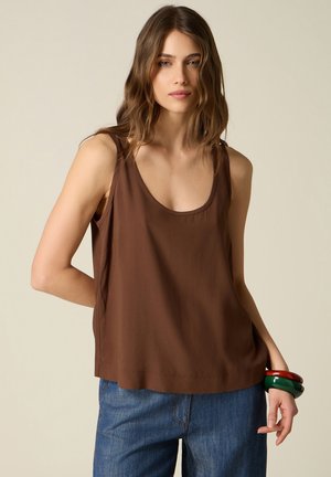 Donna con capelli castani mossi che indossa una canotta marrone, jeans blu e grossi braccialetti rossi e verdi al polso destro.