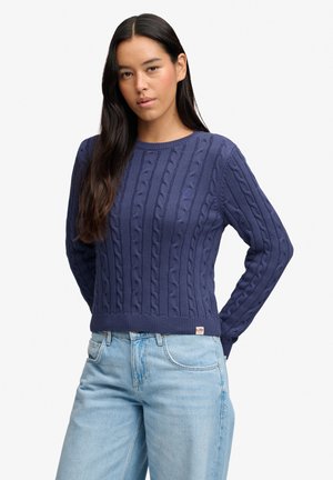 Kvinde med langt mørkt hår iført en marineblå kabelstrikket sweater og lyseblå jeans, står med hænderne bag ryggen.