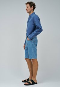 Hombre con camisa de manga larga azul, pantalones cortos de mezclilla y sandalias negras está de perfil con una mano en el bolsillo sobre un fondo gris.