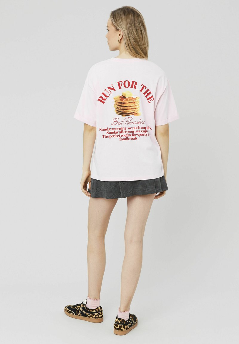 T-shirt en coton rose avec le texte "COURIR POUR LES MEILLEURES CRÊPES" et un graphique de crêpes dans le dos. Associé à une jupe plissée noire et des chaussures à imprimé léopard.