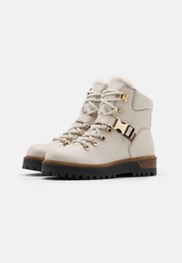 Bottes en cuir beige à la cheville avec des lacets élastiques, des garnitures en or et une doublure en fausse fourrure. Elles présentent une semelle noire épaisse et des accents en bois au talon.