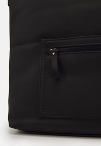 Particolare di un angolo di una borsa in pelle nera con texture, dotato di una piccola tasca con cerniera e un’etichetta in tessuto su sfondo bianco.