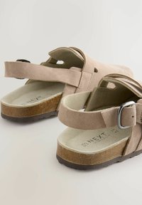 Sandales en suède beige avec un dos ouvert et une sangle réglable, dotées d'une semelle intérieure texturée et d'une semelle en caoutchouc marron.