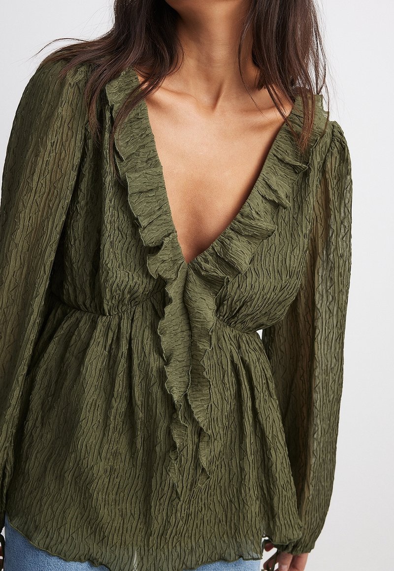 Femme portant un chemisier texturé vert olive avec un col en V profond et une bordure à volants, associé à un jean bleu clair.