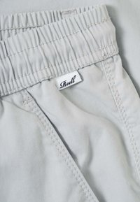 Pantalones de cintura elástica gris claro con costuras visibles y una pequeña etiqueta de tela blanca con la etiqueta "Reell" cerca de la parte superior.