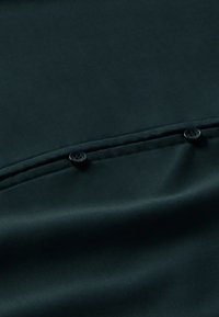 Dunkler Stoff mit einer glatten Textur, zwei schwarzen Knöpfen und einem subtilen Nahtdetail an der Oberseite.