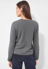 Frau mit langen braunen Haaren, die einen schlichten grauen Langarmpullover und dunkelblaue Jeans trägt und von einem weißen Hintergrund abgewandt ist.