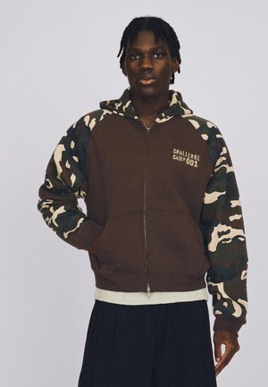 Jeune homme portant un sweat à capuche zippé marron avec des manches camouflage et le texte "Challenge Camp 001" sur la poitrine, sur un fond uni.