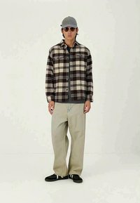 Črna in rjava karirana srajca čez sivo majico, bež široke hlače, črni teniske, siva kapa in temna sončna očala. Preprost dizajn in sproščen kroj.