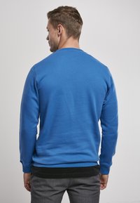 Vīriešu zils sporta krekls ar garām piedurknēm, apaļu kakla izgriezumu, ribota aploka un apakšmala; mīksta tekstūra; uzvilkts virs melna krekla.