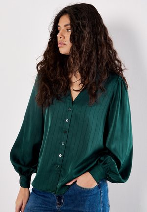 Donna con lunghi capelli ricci che indossa una camicia verde scuro con bottoni e maniche a sbuffo, insieme a jeans blu, in piedi con una mano in tasca.