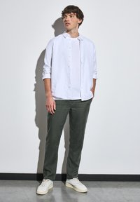 Valkoinen pellavapaitapusero, joka on kerroksittain tavanomaisen valkoisen t-paidan päällä, yhdistettynä vihreisiin housuihin ja valkoisiin tennareihin, yksinkertaista taustaa vasten.