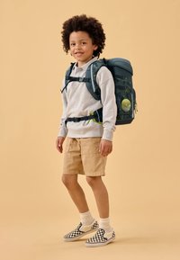 Rucksack aus dunkelgrünem Material, mit verstellbaren Trägern und lebhaftem Waldmuster. Begleitet von einem grauen Hoodie, beigen Shorts und Karo-Schuhen.