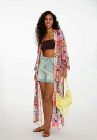 Długi, wzorzysty kimono w intensywnym różowym, niebieskim i zielonym kolorze, nałożone na brązowy crop top i jasne szorty dżinsowe. Beżowe sandały, żółta torba.