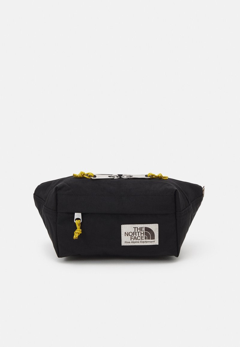 Sac en tissu noir de forme rectangulaire, avec un cordon de serrage jaune et un clip en métal argenté. Comprend une pièce de logo sur le côté.