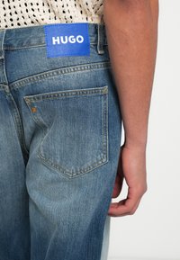 Jeans de mezclilla azul con una etiqueta de cuero con la marca "HUGO" en la cintura; presenta un ligero desgaste y dos bolsillos traseros.