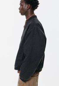 Crna bomber jakna opuštenog kroja, s rebrastim manžetama i ovratnikom. Izrađena od izdržljivog materijala s glatkom teksturom. Bočne džepove su vidljive.