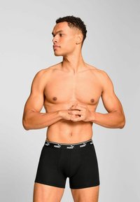 Black boxer briefs made of cotton with an elastic waistband featuring a white logo. Model stands with arms folded, showing a neutral profile.