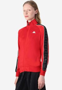 Giacca sportiva rossa con zip, caratterizzata da un colletto alto, strisce laterali nere con motivo logo e logo bianco sul petto. Tessuto morbido al tatto.