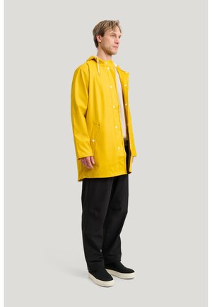 Man som bär en gul regnjacka, beige skjorta, svarta byxor och svarta slip-on skor, står i profil mot en enfärgad ljusgrå bakgrund.