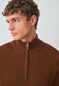 Brązowy sweter z dzianiny z ćwierć zamkiem błyskawicznym, prążkowaną teksturą, wysokim kołnierzem i małym logo na piersi. Posiada zamek błyskawiczny i gładkie wykończenie.