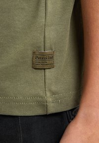 Camiseta de algodón verde oliva con un parche de logo visible, que presenta un cuello redondo, mangas cortas y un dobladillo recto con detalles de costura.