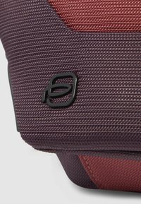 Borsa in tessuto strutturato in tonalità viola intenso e rossastre, con logo nero e design liscio e strutturato con bordi inclinati.