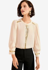 Blusa beige a maniche corte con accenti di paillettes dorate lungo le spalle, il collo e i polsini; tessuto trasparente, scollatura rotonda, vestibilità comoda.